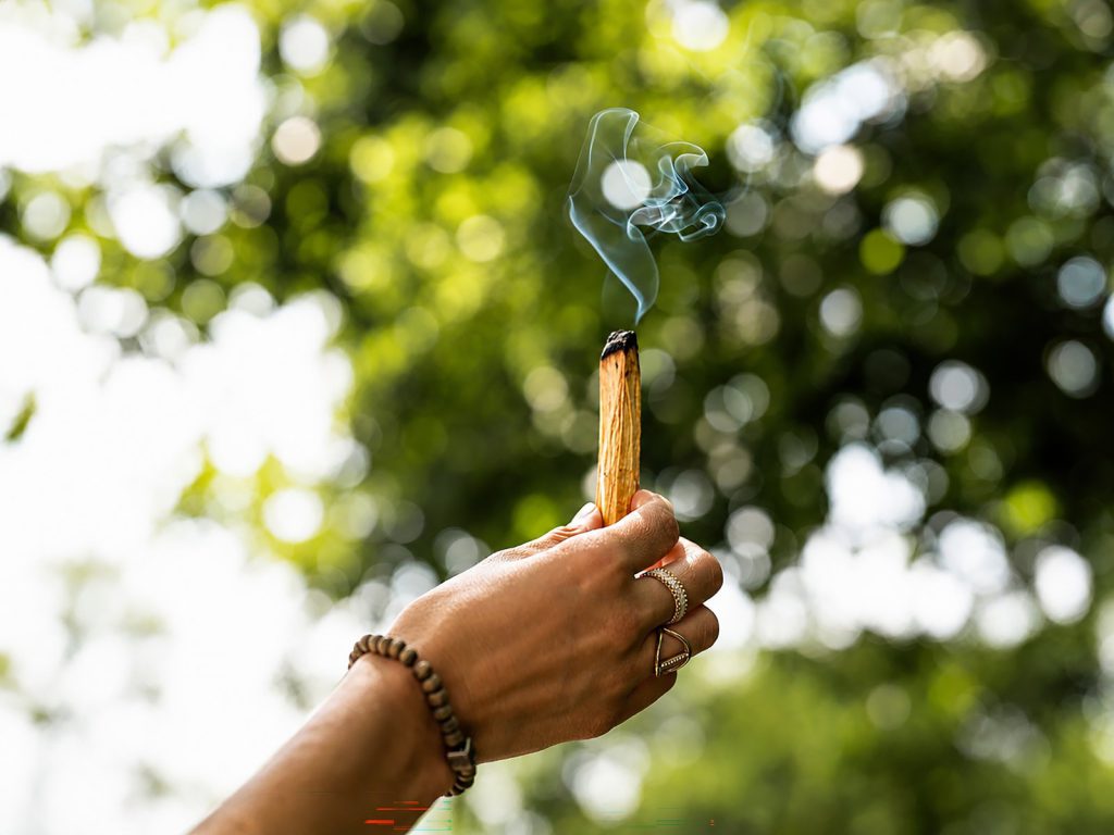 Ein glimmendes Stück Palo Santo in zwischen den Fingern einer mit Ringen und Armbändern geschmückten Menschenhand.