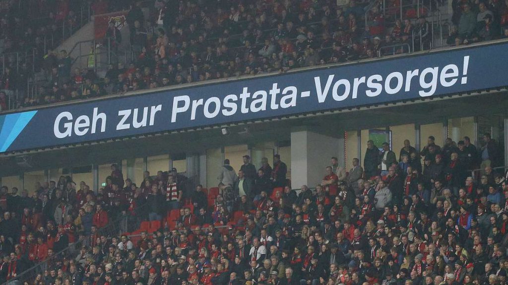 Zuschauerinnen und Zuschauer in einem Fußballstadion unter einer großen Anzeigetafel mit der Aufschrift "Geh zur Prostata-Vorsorge!"