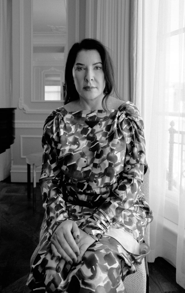 Marina Abramović sitzt in einem gemusterten Kleid an einem Fenster, Schwarz-Weiß-Porträtfotografie.