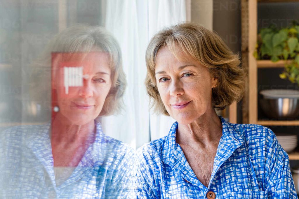 Frau in blau gemusterter Bluse steht an einem Fenster; ihr Gesicht spiegelt sich in der Scheibe.