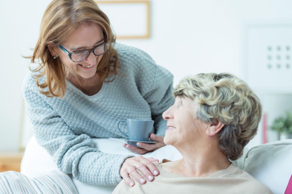 Zwei Frauen lächeln sich an; eine sitzt auf einem Sofa, während die andere ihr die Hand auf die Schulter legt und eine Tasse hält.