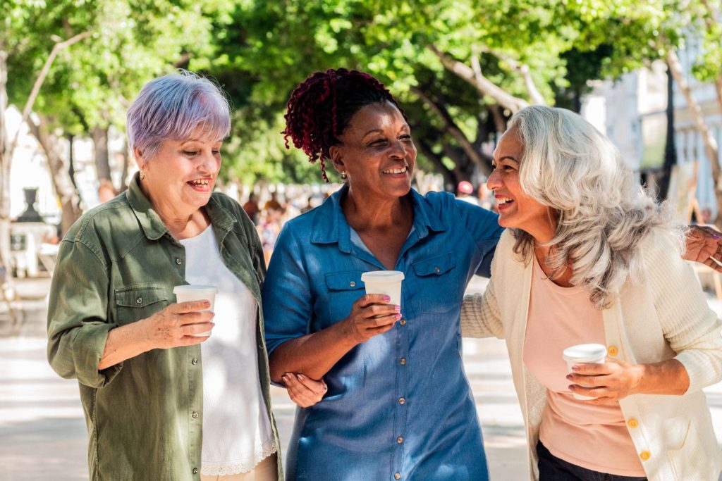 Drei ältere Frauen stehen im Freien zusammen, unterhalten sich lachend und halten Kaffeebecher in den Händen.