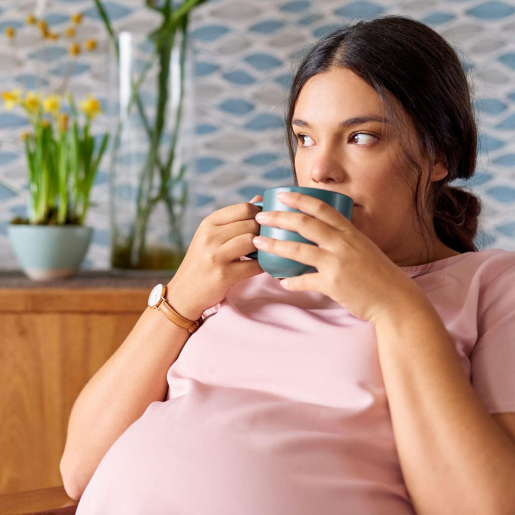 Eine schwangere Person in rosafarbenem Oberteil sitzt entspannt in einem Wohnraum und trinkt aus einer blauen Tasse. Im Hintergrund stehen gelbe Blumen und eine hohe Glasvase.