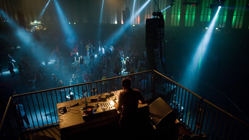 DJ legt auf einer Techno-Party in einer großen Industriehalle auf, während Besucher unter blauem Bühnenlicht auf der Tanzfläche feiern – authentische Technoszene.