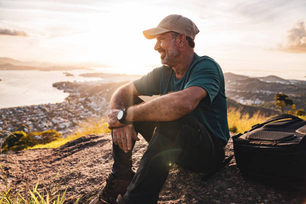 Ein Mann mit Kappe und Rucksack sitzt bei Sonnenuntergang auf einem Felsen und blickt entspannt über eine Küstenlandschaft.