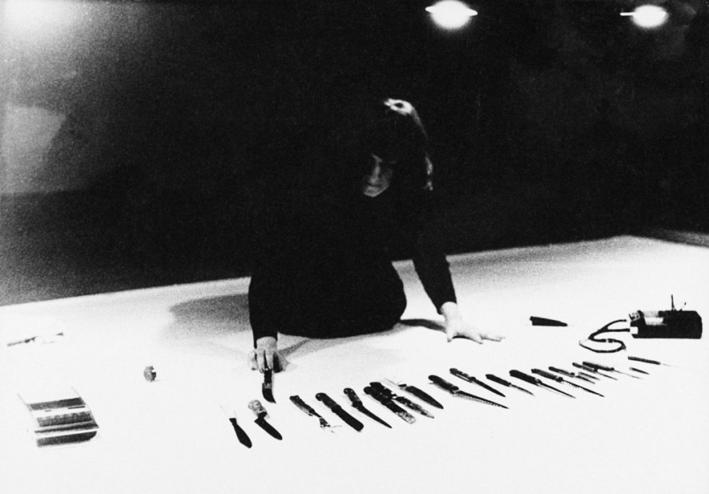 Marina Abramović bei der Performance Rhythm 10 (1973), vor einem Tisch mit einer Reihe von Messern.